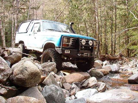 1985 Ford Bronco - A blue and white Ford Bronco is navigating a rocky stream bed in a forest. The off-road vehicle has a snorkel and auxiliary lights mounted…