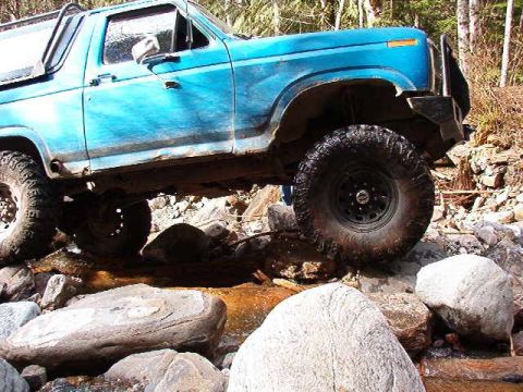 1985 Ford Bronco - The side view of a blue Ford Bronco is shown navigating a rocky stream bed. The off-road vehicle has large tires and appears to be lifted.