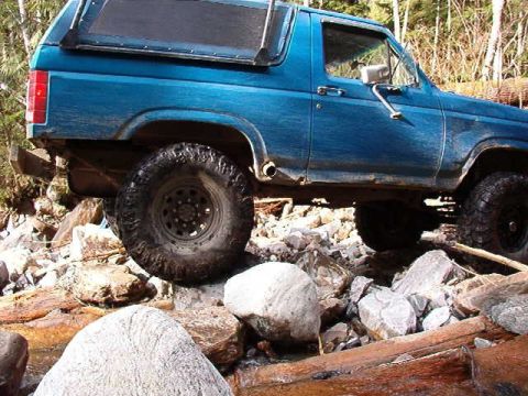 1985 Ford Bronco - A blue Ford Bronco is parked on a rocky terrain near a stream. The vehicle is angled to show its side profile, with a focus on the large of…