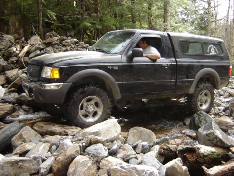 1985 Ford Bronco - A black Ford Ranger pickup truck with a camper shell navigates a rocky stream bed in a wooded area. The driver is visible through the open…