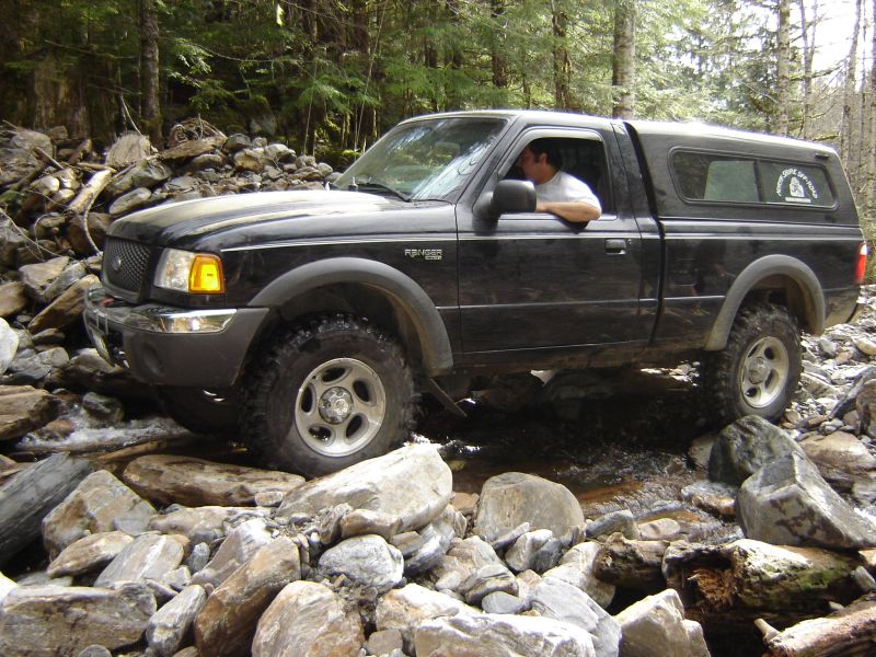 Black Ford Ranger pickup truck driving through a rocky stream in a forest.