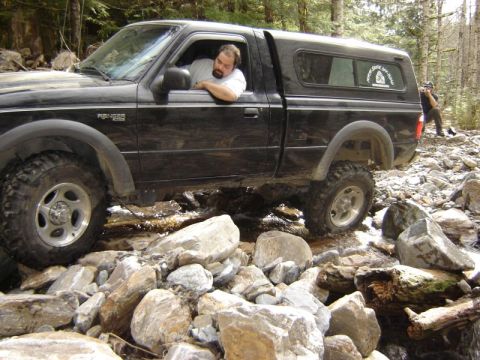 1985 Ford Bronco - A black Ford Ranger pickup truck with a camper shell navigates a rocky stream bed in a wooded area. The driver is leaning his arm on the op…