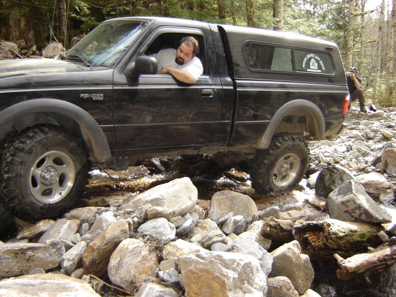 Black Ford Ranger pickup truck driving through a rocky stream in a forest.