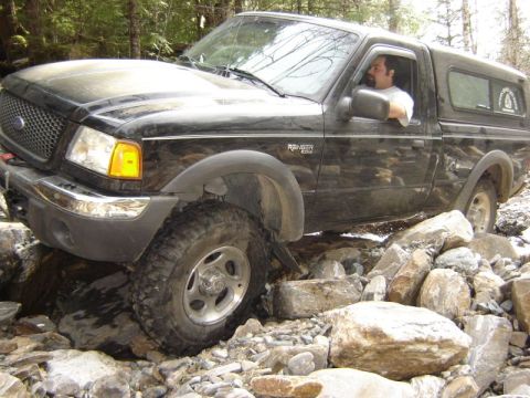 1985 Ford Bronco - A black Ford Ranger pickup truck navigates a rocky, off-road trail in a forest setting. The truck is angled, with its front driver's side t…