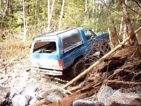 1985 Ford Bronco - A blue Ford Bronco is stuck off-road in a wooded area, leaning against fallen trees and rocks. The rear of the vehicle is visible, with its…