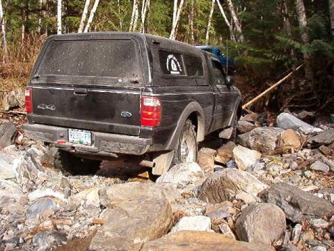 1985 Ford Bronco - A black pickup truck with a camper shell is navigating a rocky, wooded terrain. The truck's license plate reads "9230-FR".