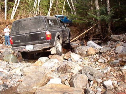 1985 Ford Bronco - A black Ford Ranger pickup truck with a camper shell navigates a rocky, off-road terrain. A person stands to the left of the truck, observi…