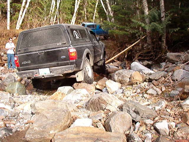 Black Ford Ranger pickup truck with camper shell driving over rocks on an off-road trail.