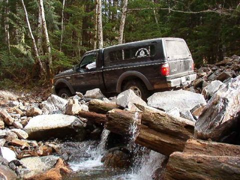 1985 Ford Bronco - A dark-colored pickup truck with a camper shell is navigating a rocky stream bed in a forest. The truck is surrounded by large rocks and fa…