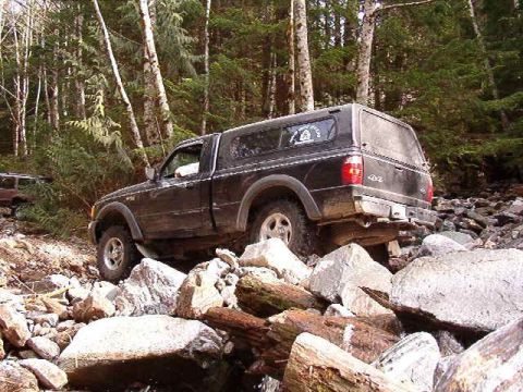 1985 Ford Bronco - A black pickup truck with a camper shell navigates a rocky, wooded trail. The truck is angled to the right, with its front wheel visible on…
