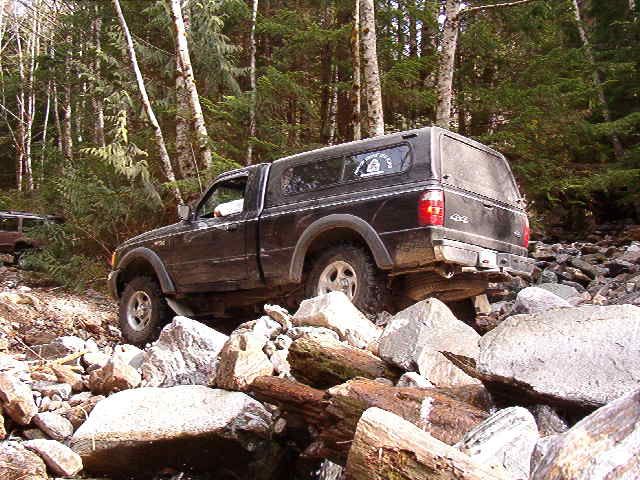 Black pickup truck with camper shell driving over rocks on a wooded trail.