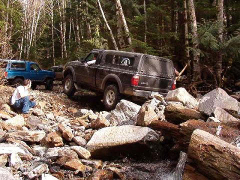 1985 Ford Bronco - A black pickup truck with a camper shell navigates a rocky, wooded trail. A blue SUV is parked behind it, and a person kneels on the rocks…