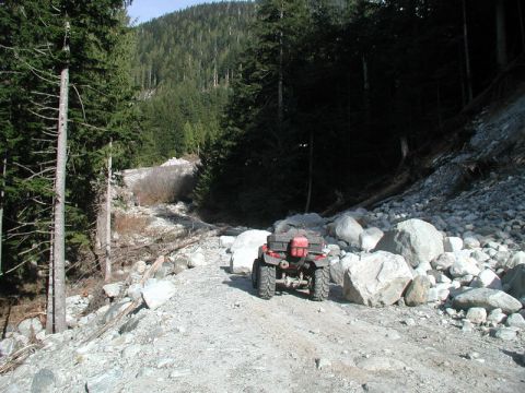1985 Ford Bronco - A red ATV with storage boxes is parked on a rocky, unpaved trail in a forested mountain area. The ATV is facing away from the camera, with…