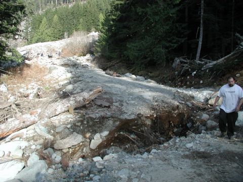 1985 Ford Bronco - A man walks on a damaged dirt road in a wooded area. The road appears to have been washed out in a section, with debris and rocks scattered…