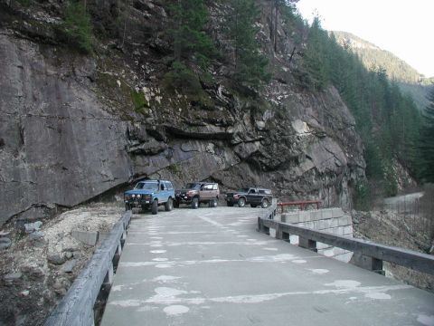1985 Ford Bronco - Three SUVs are parked on a paved surface next to a steep rock face. The vehicles are positioned on a bridge or elevated roadway with wooden…