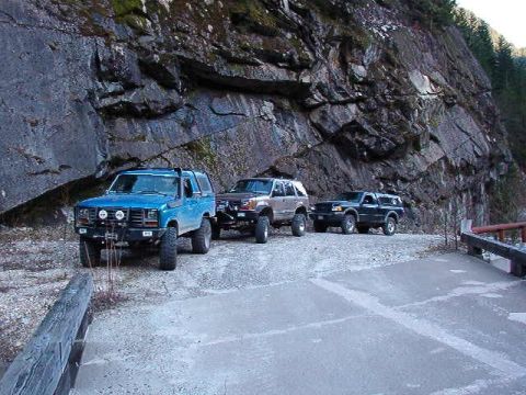 1985 Ford Bronco - A blue 1985 Ford Bronco is parked on a gravel road next to a rocky cliff face. Three off-road vehicles are visible in the scene.