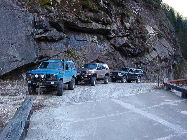 Blue 1985 Ford Bronco parked on gravel road near cliff with other off-road vehicles.