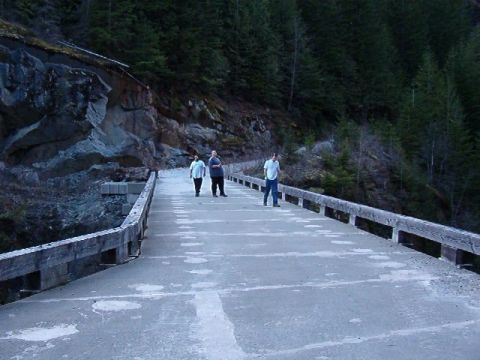 1985 Ford Bronco - Three people walk across a concrete bridge with wooden railings. The bridge is surrounded by a rocky hillside and a dense forest.