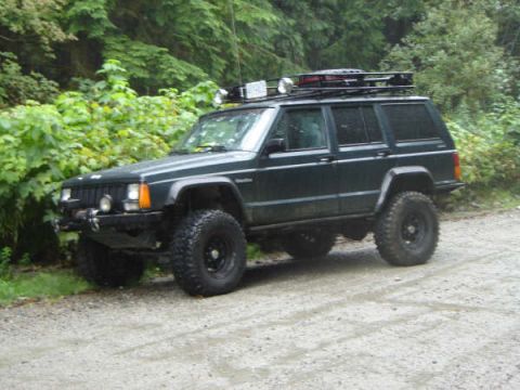1985 Ford Bronco - A dark green, lifted Jeep Cherokee XJ with off-road tires and a roof rack is parked on a dirt road next to lush green foliage.