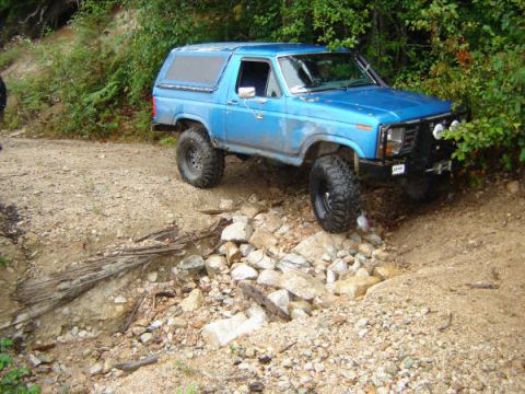 1985 Ford Bronco - A lifted, blue Ford Bronco is navigating a rocky, off-road trail with dense foliage on the right. The vehicle is angled, showing its front…