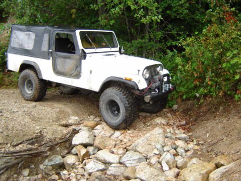 1985 Ford Bronco - A white Jeep CJ-7 with a black soft top is parked on a rocky, uneven terrain with trees and foliage in the background. The vehicle is angle…