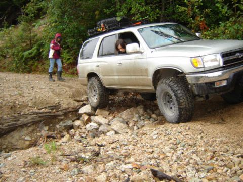 1985 Ford Bronco - A silver SUV is navigating a rocky, off-road trail with a person standing nearby. The SUV has large off-road tires and a roof rack with gea…