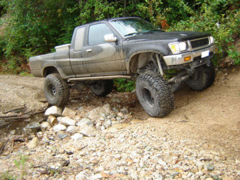1985 Ford Bronco - A lifted, dark-colored pickup truck with large off-road tires is navigating a rocky, dirt path with dense foliage in the background.