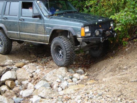 1985 Ford Bronco - A dark green Jeep Cherokee is navigating a rocky, off-road trail with dense green foliage on the right. The vehicle's front tire is angled…