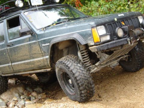 1985 Ford Bronco - A dark green, lifted Jeep Cherokee is shown at a steep angle on a dirt and rock trail. The vehicle has aggressive off-road tires, a roof ra…