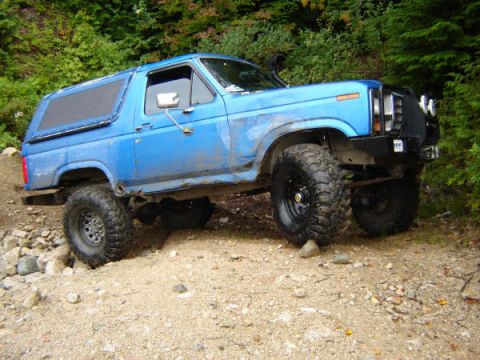 1985 Ford Bronco - A blue, lifted 1985 Ford Bronco is parked on a rocky, dirt trail surrounded by trees. The off-road vehicle has large, aggressive tires and…
