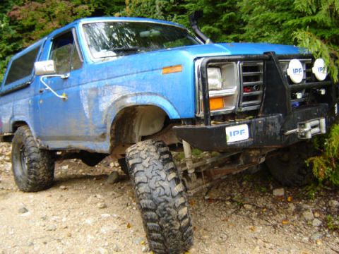 1985 Ford Bronco - A blue Ford Bronco is parked on a dirt road with trees in the background. The vehicle has a black aftermarket bumper with IPF lights and a…