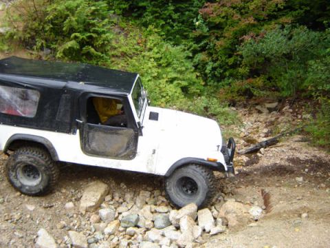 1985 Ford Bronco - A white Jeep CJ-7 with a black soft top is navigating a rocky, wooded trail. The driver's side door is open, revealing a person wearing a y…