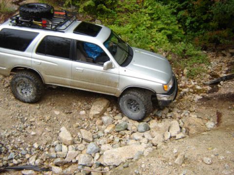 1985 Ford Bronco - A silver SUV with a roof rack is navigating a rocky, off-road trail with dense green foliage in the background.