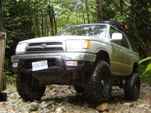 1985 Ford Bronco - A silver four-door SUV with large off-road tires is shown from a low front angle, navigating a rocky, wooded trail. The vehicle has a licen…