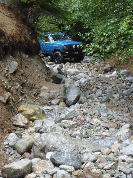 1985 Ford Bronco - A blue Ford Bronco navigates a rocky, wooded trail. The off-road vehicle is positioned at an angle, showing its front and side profile as i…