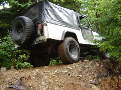 1985 Ford Bronco - A white Jeep Wrangler with a black soft top is navigating a steep, muddy incline in a wooded area. The vehicle is angled upwards, with its…