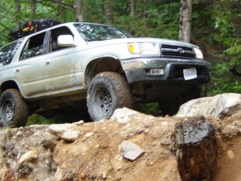1985 Ford Bronco - A silver SUV is driving on a dirt trail with large tires. The vehicle is angled to the right and is partially obscured by rocks and dirt.