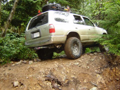1985 Ford Bronco - A silver SUV is navigating a steep, dirt trail through a dense forest. The vehicle is angled sideways, with its front tires on higher groun…