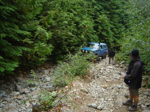 1985 Ford Bronco - A blue 1985 Ford Bronco is driving on a rocky, wooded trail. A person stands on the side of the trail looking towards the vehicle.