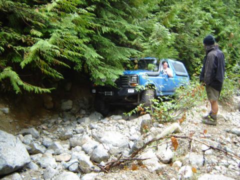 1985 Ford Bronco - A blue 1980s Ford Bronco is navigating a rocky, wooded trail with a person standing nearby watching.