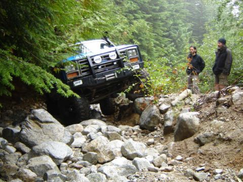 1985 Ford Bronco - A blue 1985 Ford Bronco is navigating a rocky, wooded trail. Two men stand on the side of the trail watching the off-road vehicle.