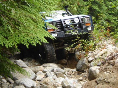 1985 Ford Bronco - A blue 1985 Ford Bronco is navigating a rocky, off-road trail, with dense green foliage obscuring part of the vehicle. The front grille and…