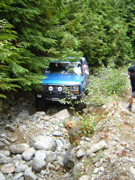 1985 Ford Bronco - A blue 1985 Ford Bronco is navigating a rocky, wooded trail. The driver is visible through the open window, and the vehicle is partially ob…