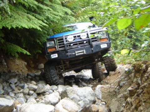 1985 Ford Bronco - A blue 1985 Ford Bronco is navigating a rocky, wooded trail, with its front tire angled upwards on a large rock.