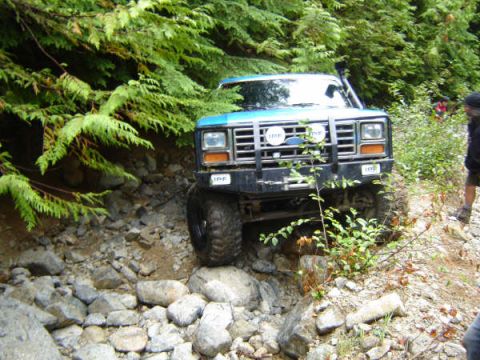 1985 Ford Bronco - A blue Ford Bronco is navigating a rocky, wooded trail. The front of the vehicle is visible, showing its grille, headlights, and large off-…