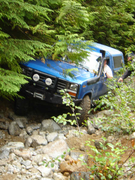 1985 Ford Bronco - A blue 1985 Ford Bronco is navigating a rocky, wooded trail, with its front tire climbing over large stones. The vehicle features a snorkel…