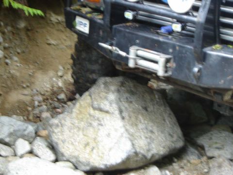 1985 Ford Bronco - A large rock is wedged under the front bumper and tire of a black off-road vehicle. The vehicle has a winch mounted on the bumper and auxil…