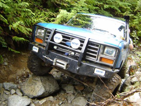 1985 Ford Bronco - A blue 1980s Ford Bronco is navigating a rocky, wooded trail, with its front tire perched on a large boulder. The vehicle features a winch…