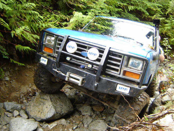 Blue 1980s Ford Bronco off-roading on a rocky trail, front tire on a boulder.