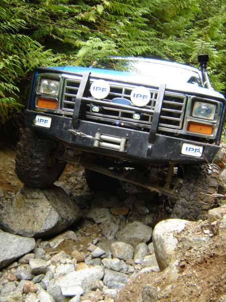 1985 Ford Bronco - A blue Ford Bronco is navigating a rocky, off-road trail with dense green foliage in the background. The front of the vehicle is angled tow…